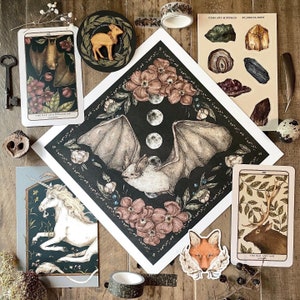 May include: A collection of art prints and stickers featuring a central square illustration of a bat surrounded by moon phases and flowers, accompanied by tarot-style cards of a bat, a unicorn, and an elk, plus a fox sticker and a gems sheet.
