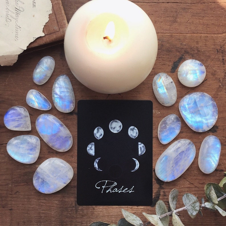 May include: A black card with the word "Phases" written in white cursive script.  The card has a circle of eight white moon phases around a black center.  The card is surrounded by a variety of iridescent moonstone crystals on a wooden surface.
