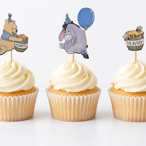 May include: Three cupcakes topped with white frosting and decorative paper toppers. One features a yellow bear with a blue party hat holding a honey pot labeled HUNNY, another shows a gray donkey with a blue balloon, and the third a honey pot.