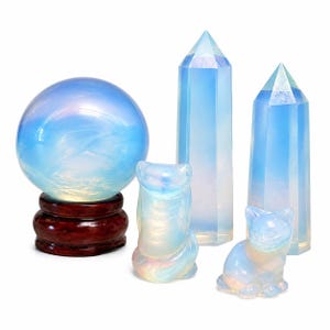 May include: A collection of opalite objects including a large smooth sphere resting on a dark wooden stand, two tall hexagonal crystal points with pointed tops, and two small carved animal figurines, one shaped like an owl and one like a cat.