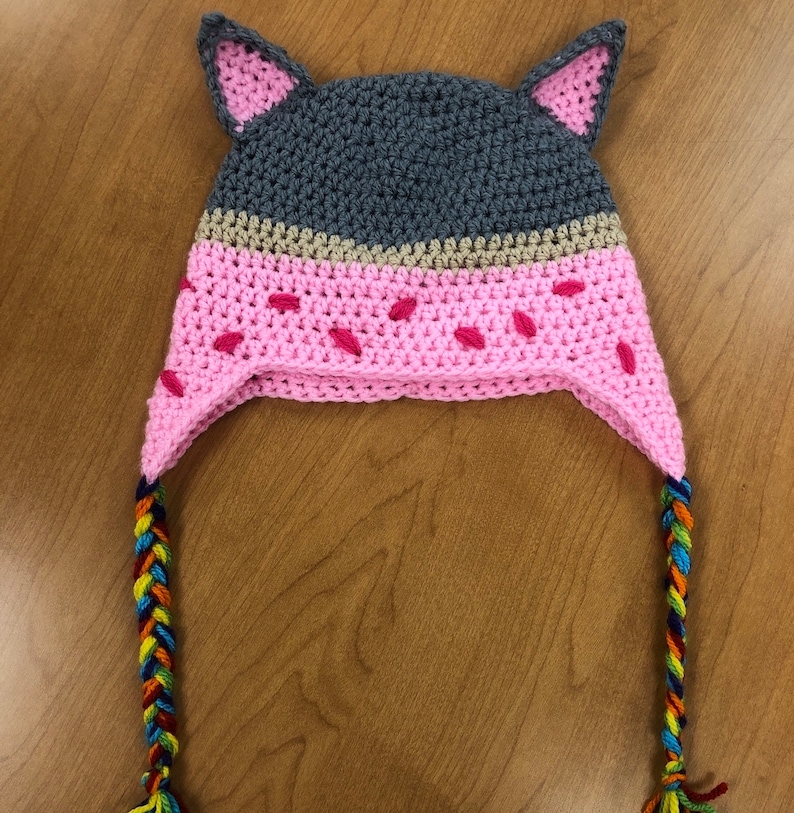 May include: A pink and gray crocheted hat with cat ears and a multi-colored braided tassel. The hat has a pink and white striped pattern.