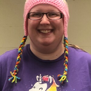 May include: A woman wearing a pink and grey crocheted hat with rainbow yarn braids. The hat has cat ears. She is wearing a purple t-shirt with a white unicorn graphic and rainbow mane.
