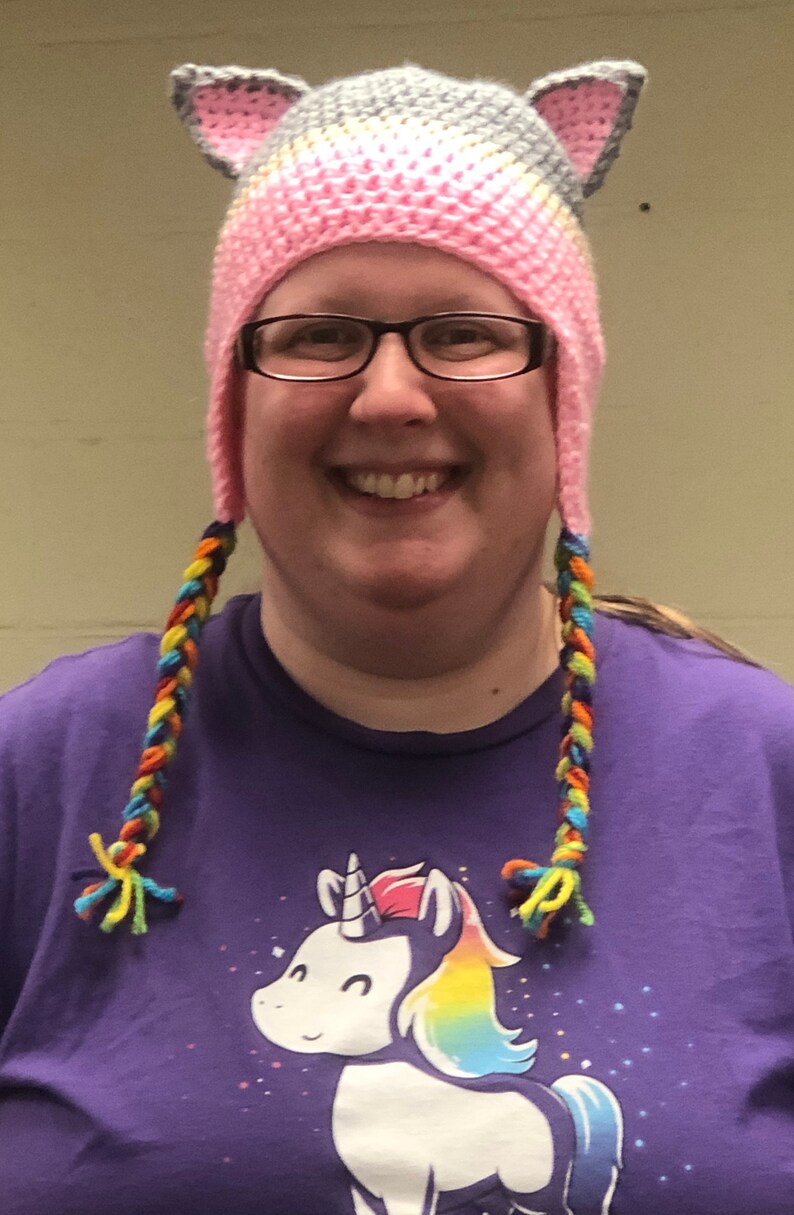May include: A woman wearing a pink and grey crocheted hat with rainbow yarn braids. The hat has cat ears. She is wearing a purple t-shirt with a white unicorn graphic and rainbow mane.