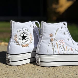 May include: White high-top Converse sneakers with black soles. The left shoe features a floral wreath with the text "Chuck Taylor All Star" and initials. The right shoe has embroidered flowers and foliage.