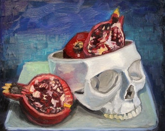 May include: A white skull with a red pomegranate inside. The skull is sitting on a blue and green surface. There is another pomegranate on the surface next to the skull.
