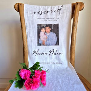 May include: White chair with a white fabric cover. The fabric has a black inscription that reads "reserved" and "In loving memory of the father of the groom, Marvin Deleon, watching us from heaven." A photo of a man and a young girl is in the centre of the fabric. A bouquet of pink flowers is in the foreground.