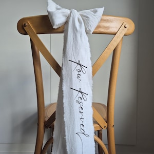 Könnte beinhalten: Ein weißer Stoff-Stuhlschleife mit einer schwarzen Schleife und dem Text "Your Reserved" in Kursivschrift.