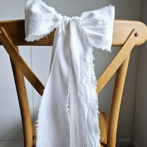 Op de afbeelding: Een witte stoffen strik gebonden om de rugleuning van een houten stoel. De strik is gescheurd en heeft een rustieke uitstraling.