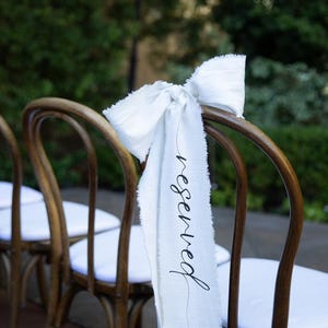 May include: A white fabric ribbon tied in a bow on the back of a wooden chair. The ribbon has the word "reserved" written on it in black calligraphy.