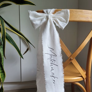 May include: White chair back with a white fabric bow tied around it. The fabric has the word "Matchmaker" written on it in black ink.