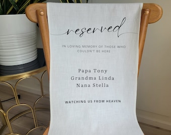 May include: A white chair with a fabric sash that reads "reserved" in black script. The sash also includes the text "In loving memory of those who couldn't be here" and lists the names "Papa Tony, Grandma Linda, Nana Stella" followed by "Watching us from heaven."