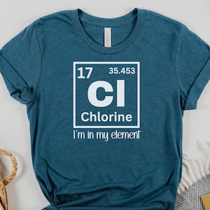 Puede incluir: Una camiseta azul turquesa con un gráfico blanco del elemento cloro de la tabla periódica. El texto "Estoy en mi elemento" está impreso debajo del gráfico.