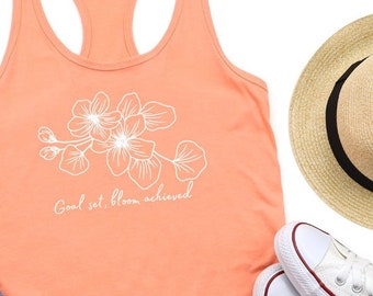 Könnte beinhalten: Ein pfirsichfarbenes Tanktop mit einem weißen Blumenmuster und dem Text "Goal set. Bloom achieved." Das Tanktop liegt flach auf einem weißen Hintergrund mit einer Jeans, einem Strohhut und weißen Turnschuhen.