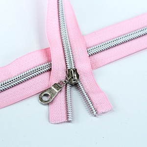 May include: A pink zipper with a silver slider and teeth. The zipper is open and the slider is in the middle of the zipper.