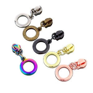 May include: A collection of metal zipper pulls in various colors, including silver, bronze, black, gold, rose gold, and a rainbow iridescent finish. Each pull features a circular ring and a zipper head.