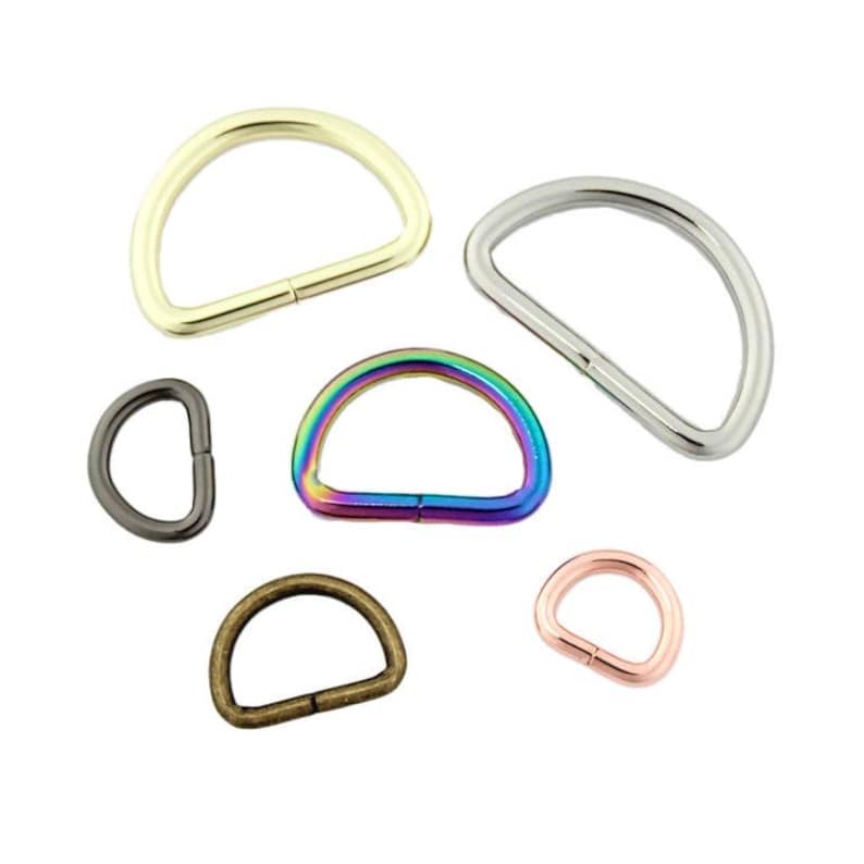 May include: A collection of six metal D-rings in various finishes including gold, silver, iridescent rainbow, gunmetal, antique bronze, and rose gold. Each D-ring features a smooth, curved top and a flat base, designed for strap attachment.