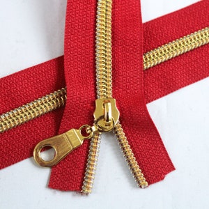 May include: Red zipper tape with gold metal teeth and a gold pull tab. The zipper is open and the two sides are separated.