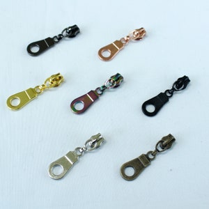 May include: Assortment of six zipper pulls in various colours and finishes, including gold, silver, black, and iridescent. The zipper pulls are all metal and have a simple, functional design.