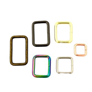 May include: A collection of six rectangular metal loops in various finishes including antique brass, matte black, polished gold, iridescent rainbow, silver, and rose gold. Each loop features rounded corners and a smooth, continuous frame.