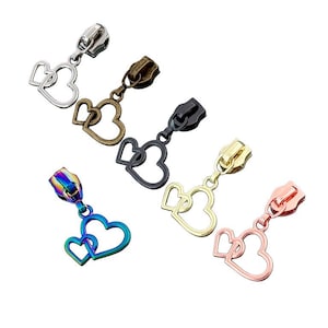 May include: A collection of zipper pulls in various metallic finishes, including silver, bronze, black, gold, iridescent, and rose gold. Each pull features a decorative double heart charm. These zipper pulls are ideal for adding a personalized touch to garments and accessories.