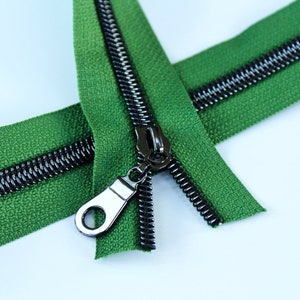 May include: A green zipper with a black metal slider and teeth. The zipper is open and the slider is in the middle of the zipper.
