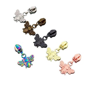 May include: A collection of six metal zipper pulls, each featuring a bee-shaped charm. The set includes finishes in silver, antique bronze, matte black, iridescent rainbow, gold, and rose gold, all attached to standard zipper slider mechanisms.