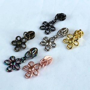 May include: Assortment of six metal zipper pulls in various colors and finishes. Each pull features a flower design with a loop for attaching to a zipper.