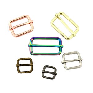 May include: A collection of six rectangular metal strap adjusters in various finishes including rose gold, gold, iridescent rainbow, antique bronze, gunmetal, and silver, each featuring a central sliding bar for webbing adjustment.