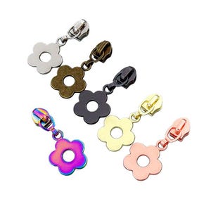 May include: A collection of flower-shaped zipper pulls in various metallic finishes. The pulls include silver, bronze, black, gold, rose gold, and a rainbow iridescent finish. Each pull has a small, round hole in the center of the flower.