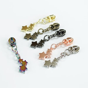 May include: Five zipper pulls with star-shaped charms. The charms are in different colours, including gold, silver, rose gold, black, and rainbow.