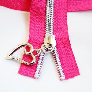 May include: A close-up of a pink zipper with a silver heart-shaped pull. The zipper is open and the teeth are visible.