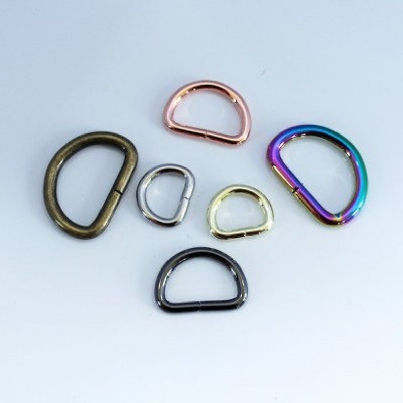 May include: A collection of six metal D-rings in various finishes and sizes. The set includes a large antique brass ring, a rose gold ring, a small silver ring, a small gold ring, a medium black ring, and a medium iridescent rainbow ring.