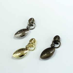 May include: Three zipper pulls in different finishes: silver, gold, and bronze. Each pull features a football-shaped charm.