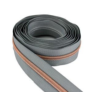 May include: A roll of gray zipper tape with a copper-colored zipper. The zipper is coiled, showing the length of the tape. The tape is a solid gray color, and the zipper teeth are a contrasting copper color.