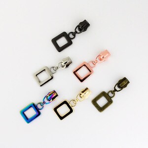 May include: Five zipper pulls with square-shaped charms. The charms are in different colours: black, silver, rose gold, gold, and iridescent.