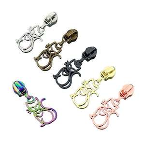 May include: A collection of five metal zipper pulls, each shaped like a stylized cat with a heart cutout in the center. The set includes finishes in silver, antique bronze, matte black, gold, and a multi-colored iridescent rainbow tone.
