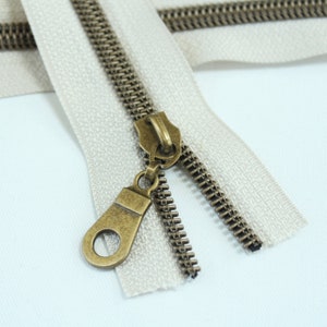 May include: A close-up of a beige zipper with a brass slider and pull. The zipper is open and the slider is in the middle of the zipper tape.