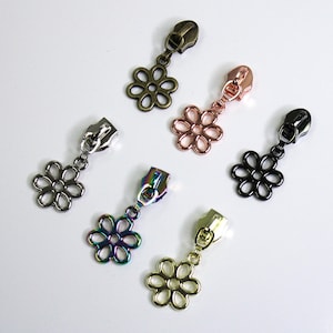 May include: Six zipper pulls with flower-shaped charms. The charms are in different colors, including gold, silver, black, rose gold, and iridescent.