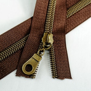 May include: A close-up of a brown zipper with antique gold metal teeth and a slider. The zipper is open and the slider is in the middle of the zipper.