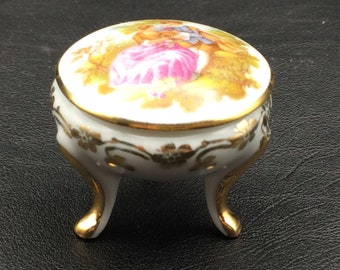 May include: A small, white porcelain trinket box with gold accents and a painted floral design. The lid features a painted scene of two figures in a garden setting.