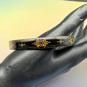 Peut inclure: Un bracelet jonc noir avec un motif floral décoratif en or et blanc. Le bracelet est tenu sur une main noire, mettant en valeur le design complexe et le contraste des couleurs. Le bracelet est un bijou.
