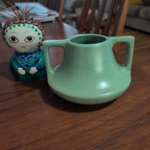 Puede incluir: Un recipiente de cerámica verde claro mate con dos asas laterales y una base ancha, junto a una pequeña figura de cerámica pintada a mano con un vestido verde azulado y una planta de aire espinosa sobre la cabeza.