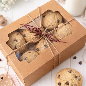 May include: A kraft paper box tied with twine, filled with chocolate chip cookies. The box has a clear window, showcasing the cookies. A single cookie sits beside the box, with a bottle of milk in the background. The cookies are golden brown with chocolate chips.