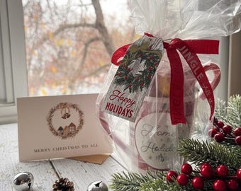May include: A holiday gift set with a red and white plaid mug, a gift tag that says "Happy Holidays", and a greeting card that says "Merry Christmas to All". The set is wrapped in clear cellophane and tied with a red ribbon. Silver bells and pinecones are scattered around the set.