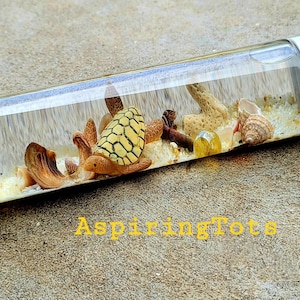 May include: A clear plastic cylinder filled with white sand, seashells, a small starfish, and a brown and yellow turtle figurine. The cylinder has a white cap.