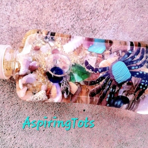 May include: A clear glass bottle filled with sand, seashells, and a small blue glass pipe. A black and blue crab is visible in the bottle.