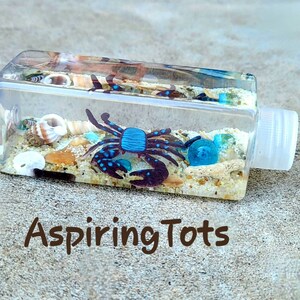 May include: A clear plastic bottle filled with sand, seashells, and a blue and black plastic crab. The bottle has a white cap and the text "Aspiring Tots" is printed on the surface below the bottle.
