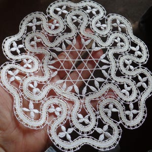 Peut inclure: Un napperon en dentelle blanche fait à la main avec un motif floral et géométrique. Le napperon présente un motif central en forme d'éclat d'étoile entouré de motifs floraux interconnectés et d'un bord festonné. La dentelle délicate est tenue dans une main.