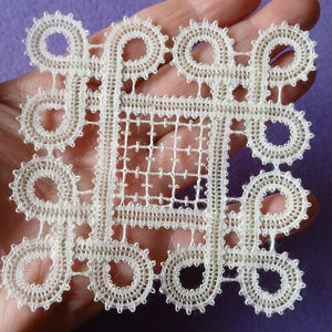 Può includere: Un centrino in pizzo quadrato color bianco sporco caratterizzato da un intricato motivo a nodi con bordi ad anello e un design centrale a griglia. Il delicato ricamo mostra un motivo geometrico ripetitivo con dettagli in filo fine.