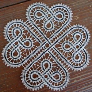 Op de afbeelding: Een handgemaakte witte kanten doily met een klaverbladontwerp. Het ingewikkelde patroon heeft een herhalend motief van onderling verbonden harten en lussen, met zwarte accenten. De doily ligt op een houten oppervlak.